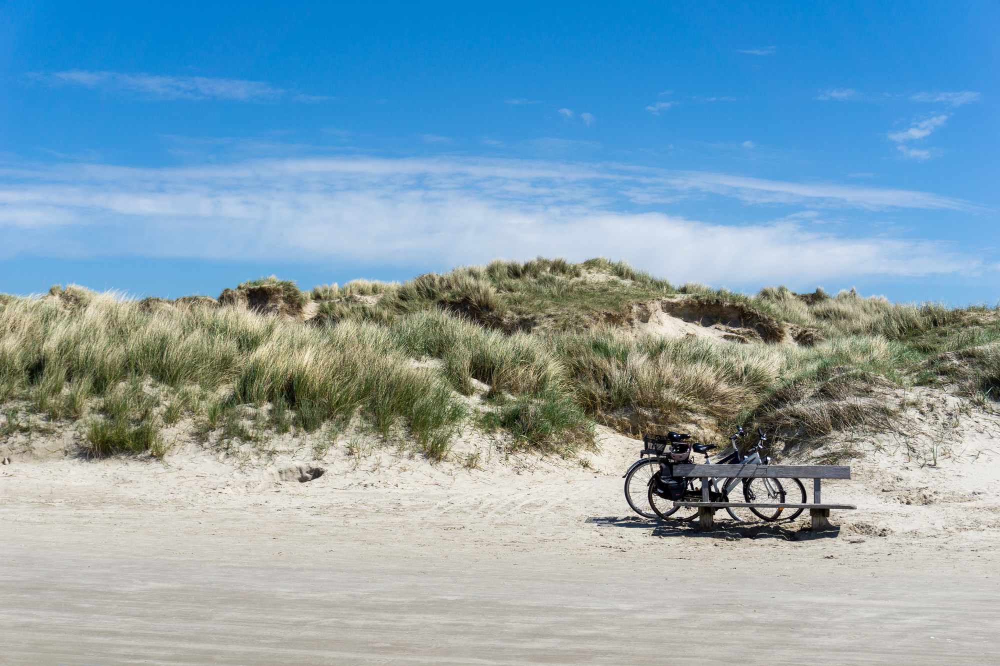 Zwei Fahrräder, die vor einer großen Sanddüne mit Gräsern und Schilf unter blauem Himmel abgestellt sind